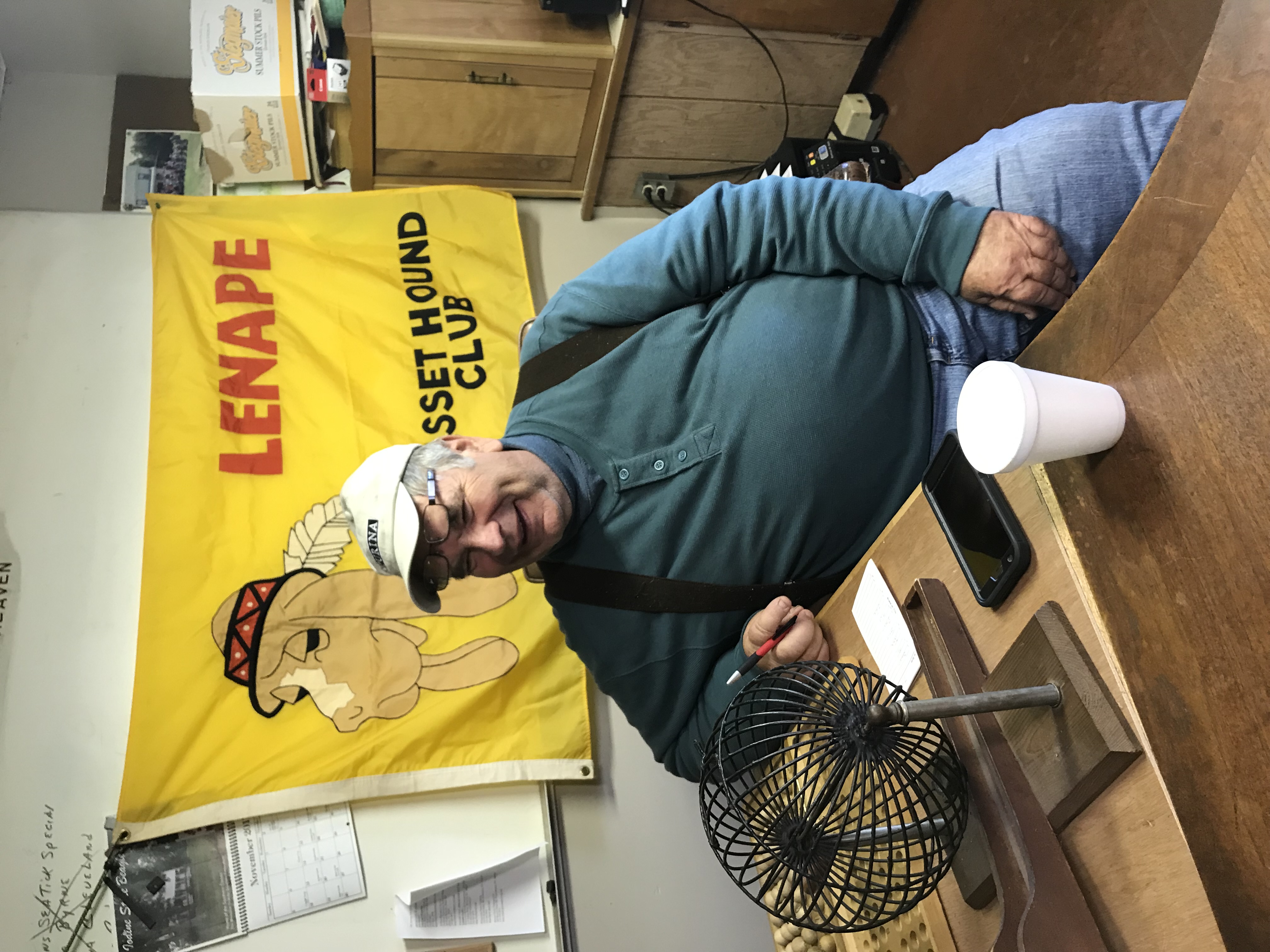 a man sits in front of the Lenape Basset Hound Club flat, near a bingo ball roller used to brace the hounds