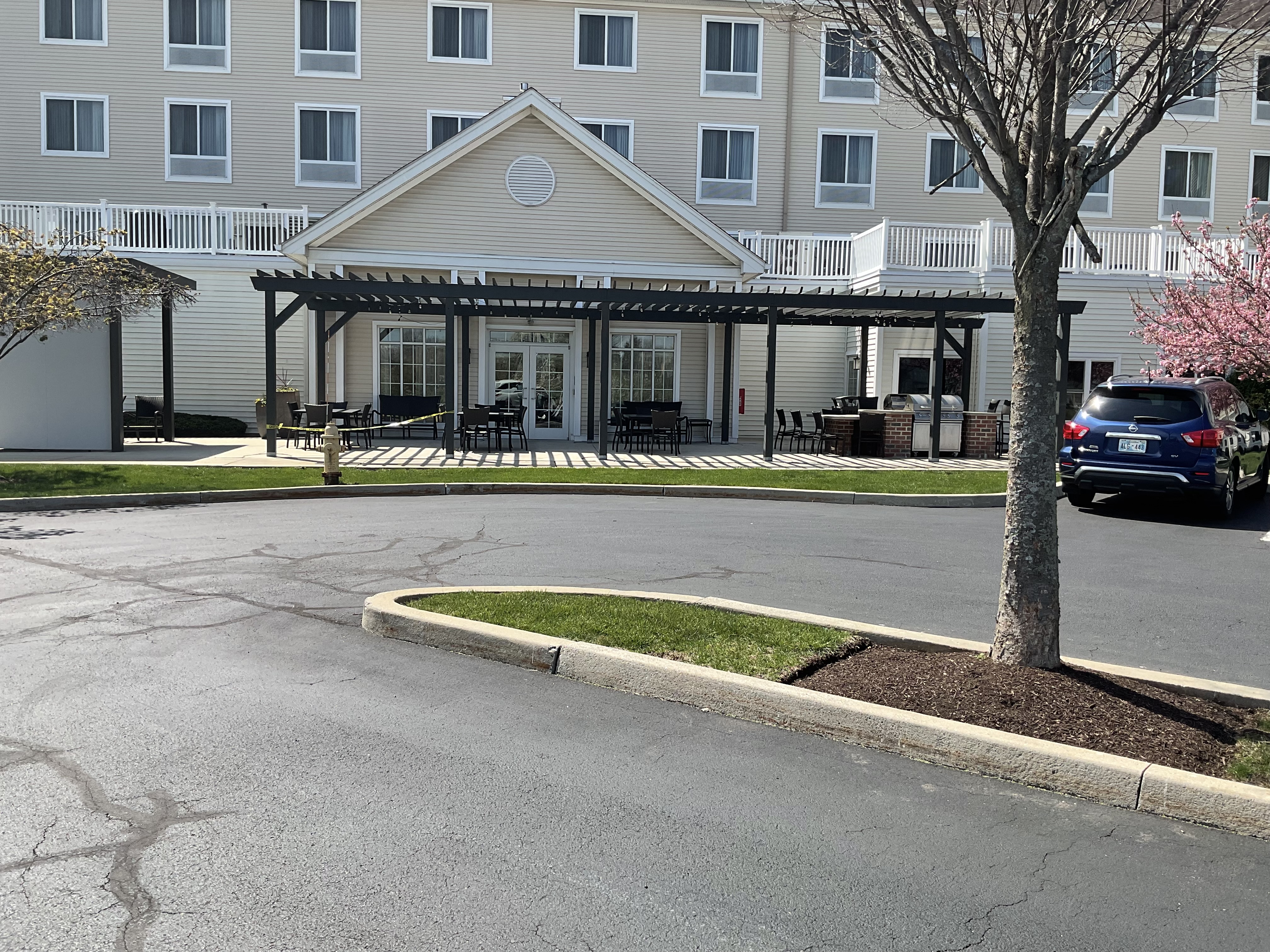 Rear of a hotel building that has a grassy area and a pergola. Parking lots with an island with a tree.