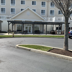 Rear of a hotel building that has a grassy area and a pergola. Parking lots with an island with a tree.