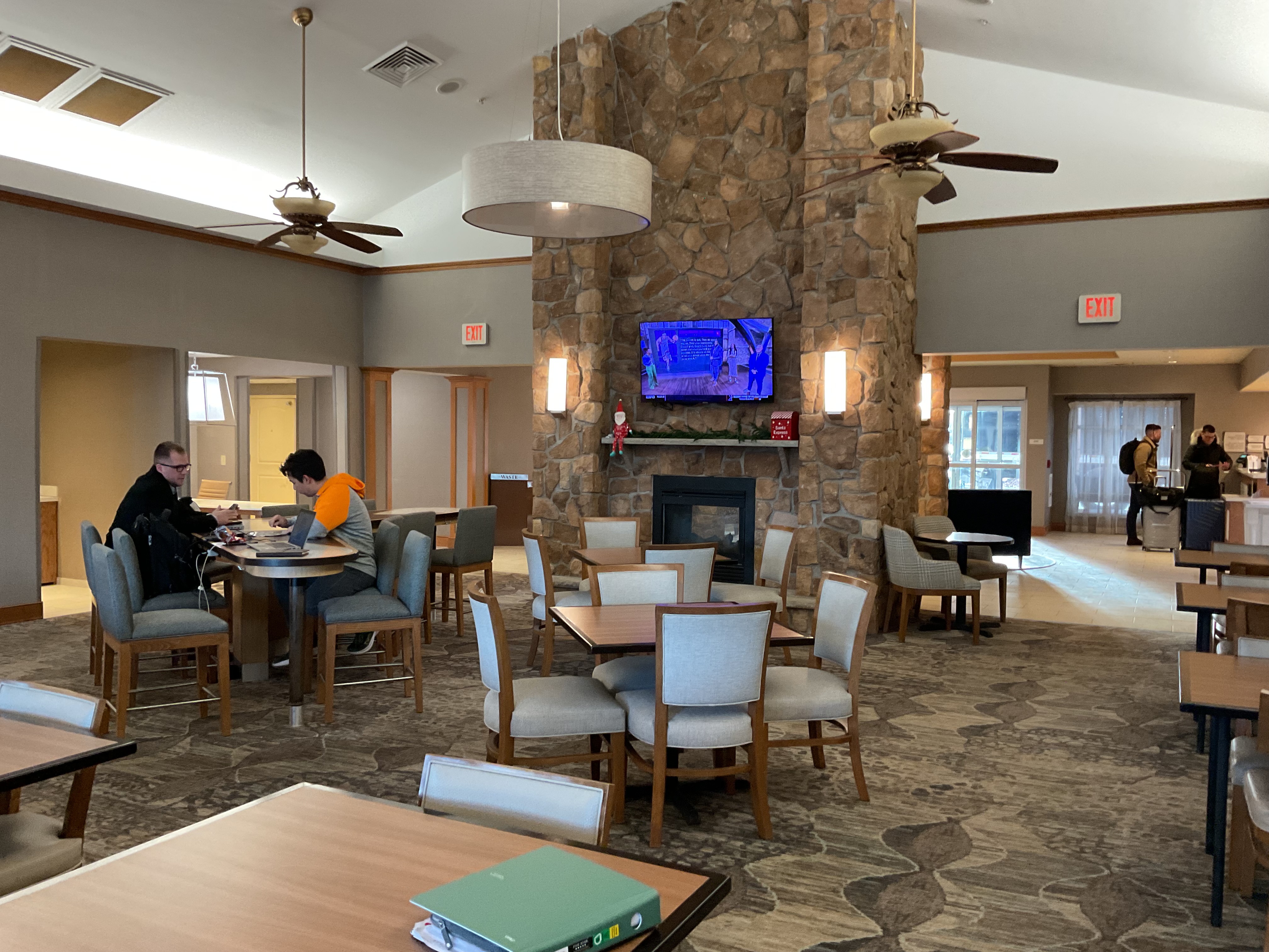 Inside of hotel shows a large fireplace in the center of the room with stonework going to the ceiling. Multiple sets of tables with chairs and a view of the front desk for checking in the background.