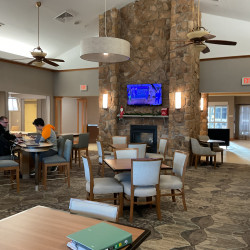 Inside of hotel shows a large fireplace in the center of the room with stonework going to the ceiling. Multiple sets of tables with chairs and a view of the front desk for checking in the background.