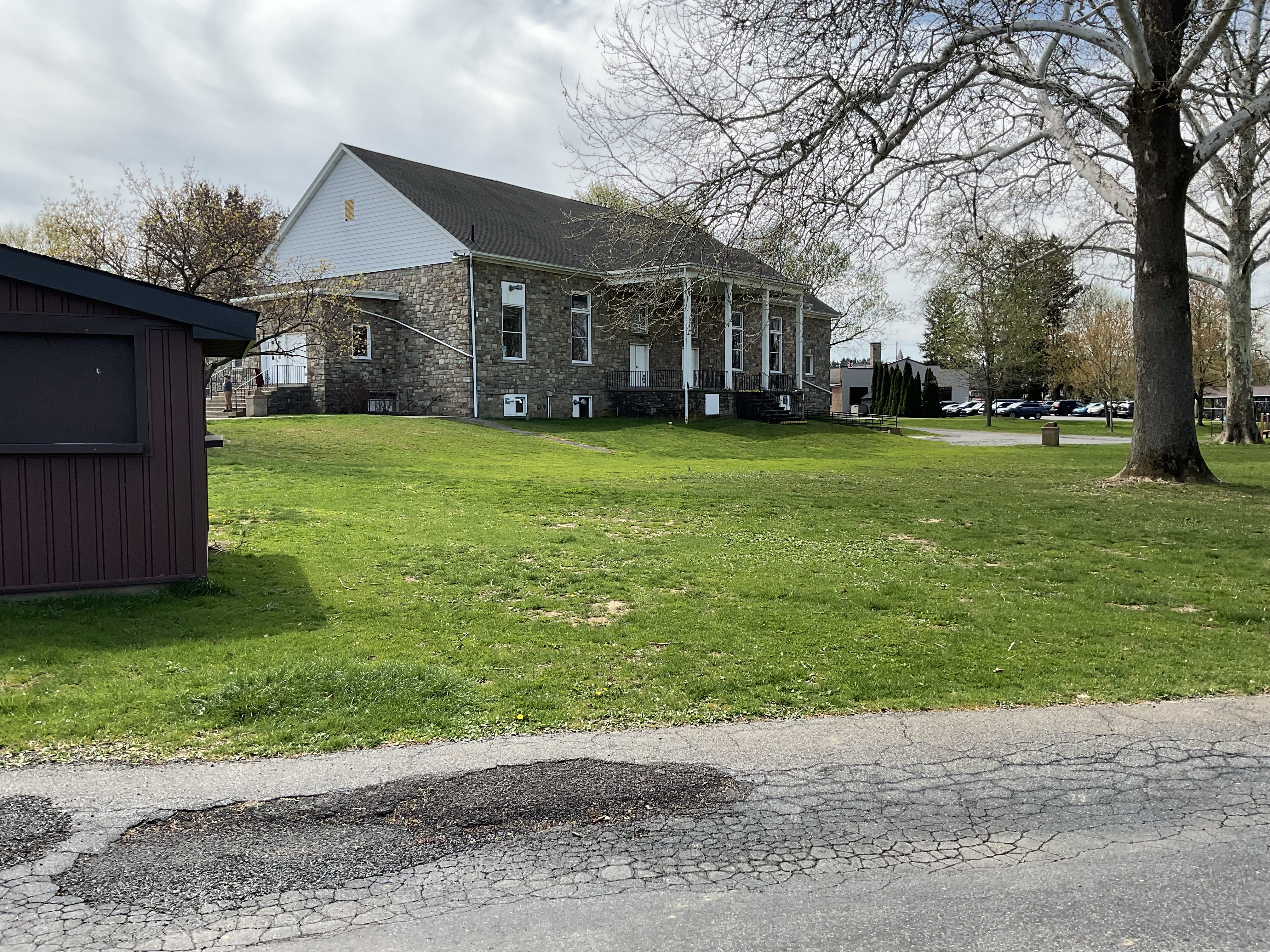 Building in center of picture with first story partially below ground. Main story has steps leading up to it. Trees on the right side and grassy area in front of the building. Also shows a paved driveway in the foreground.