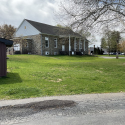 Building in center of picture with first story partially below ground. Main story has steps leading up to it. Trees on the right side and grassy area in front of the building. Also shows a paved driveway in the foreground.
